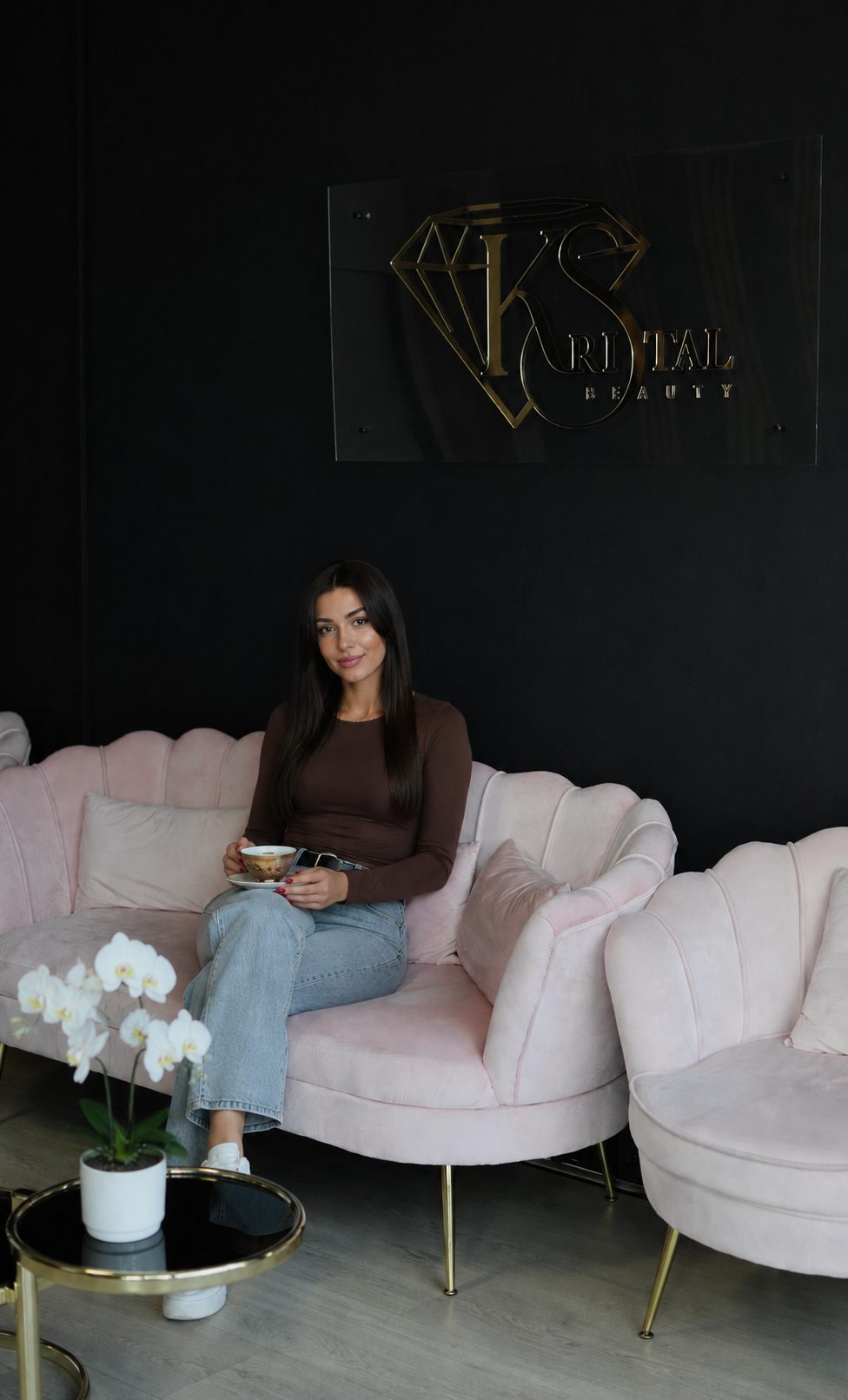 Een bezoekster met een kopje op de roze schelpsofa, onder het Kristal Beauty-sign