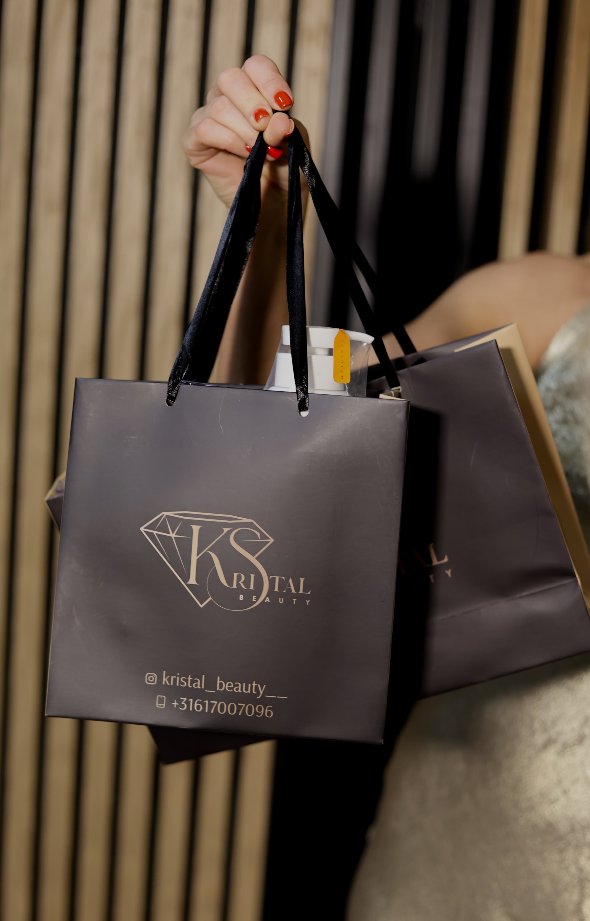 Twee donkere Kristal Beauty-tasjes met het KS-logo, omhoog gehouden voor een houten lamellenwand